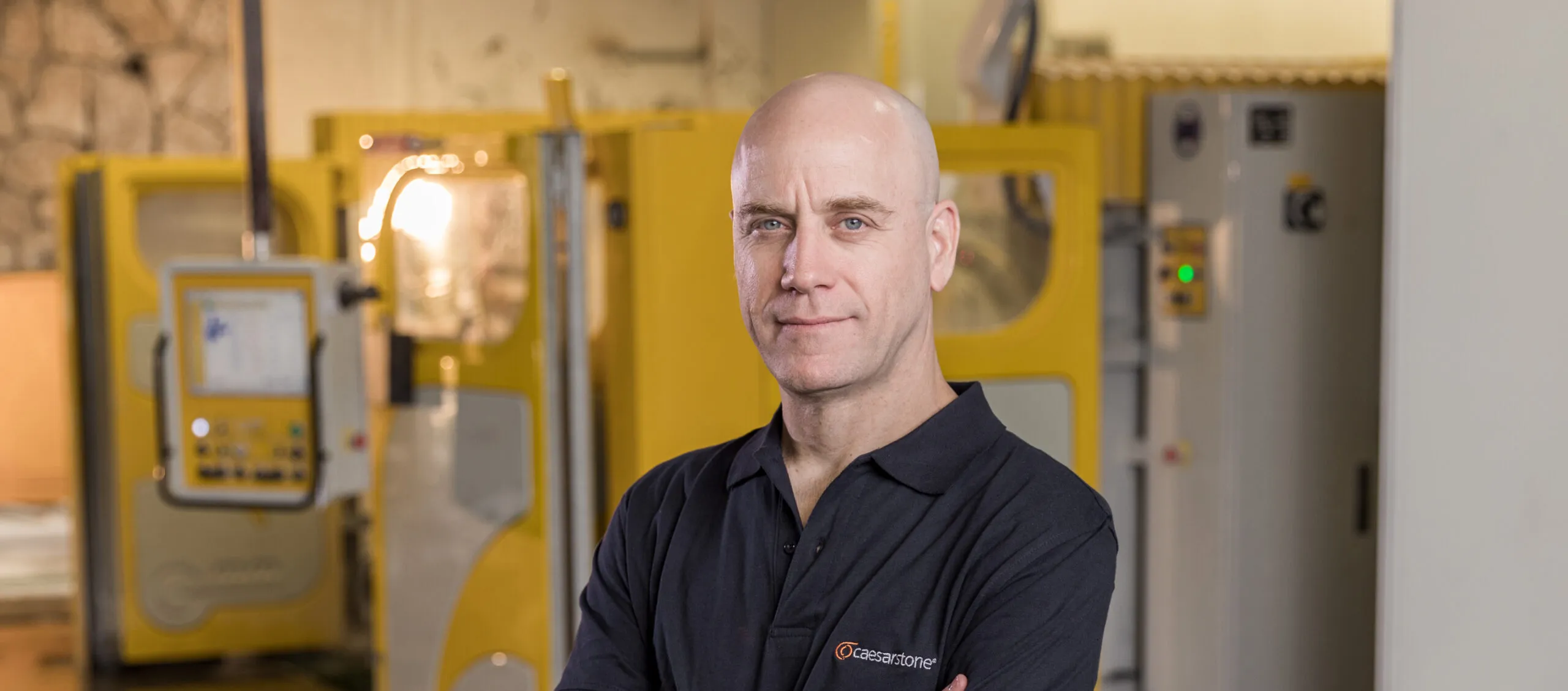Person wearing a Caesarstone shirt standing with folded arms in a fabrication workshop, showcasing professionalism and expertise.