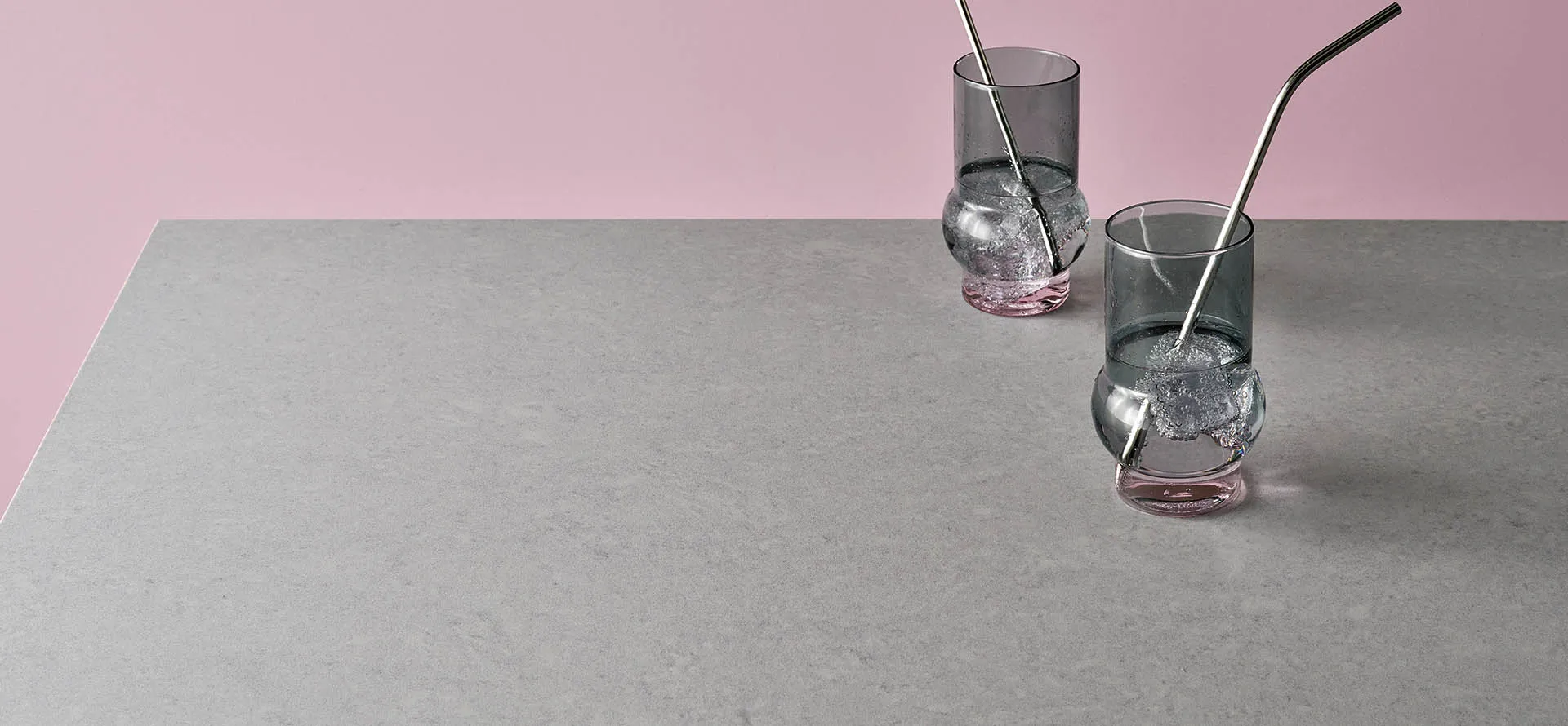 Grey Caesarstone quartz countertop with two glasses of sparkling water and metal straws on a pink background.