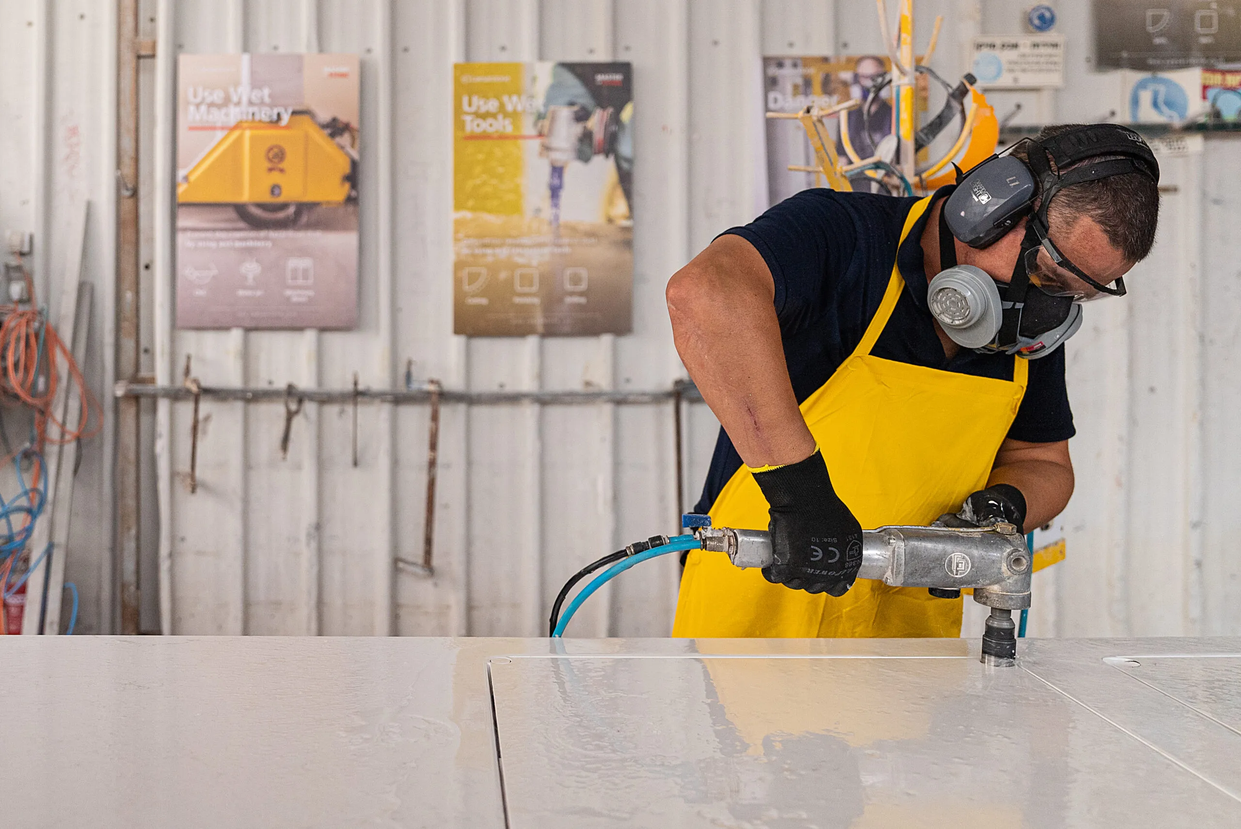 Worker wearing protective gear and yellow apron uses power tool on a white surface.